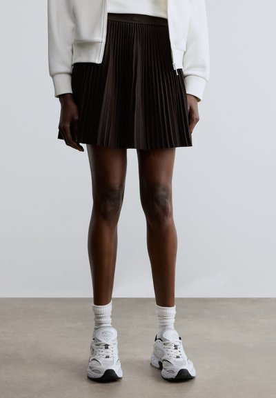 Jupe plissée marron foncé en tissu, associée à des baskets blanches et des chaussettes côtelées. La jupe présente une taille haute et un design fluide.