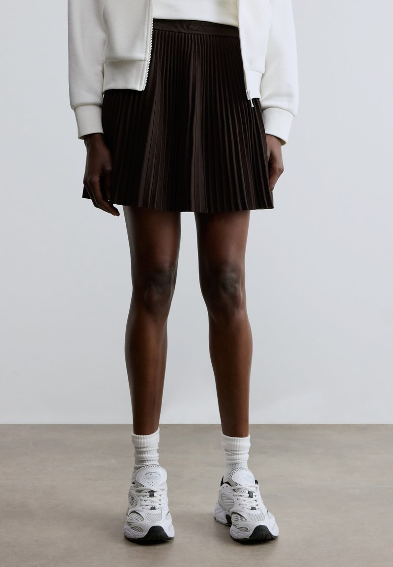 Jupe plissée marron foncé en tissu, associée à des baskets blanches et des chaussettes côtelées. La jupe présente une taille haute et un design fluide.