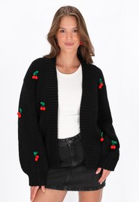 Juodas megztinis su raštu, turintis per didelius rankovės, su raudonų vyšnių siuvinėjimo akcentais priekyje ir rankovėse.