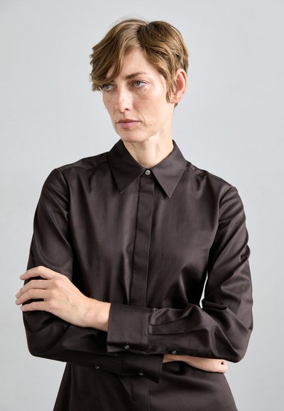 Persona con capelli corti marrone chiaro che indossa una camicia nera a maniche lunghe con bottoni, braccia incrociate, che guarda di lato su uno sfondo grigio uniforme.