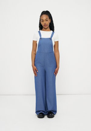 Νεαρή γυναίκα όρθια, φοράει μπλε ευρύχωρα τζιν ολόσωμα παντελόνια πάνω από λευκό κοντομάνικο τοπ με μαύρα παπούτσια, ουδέτερο φόντο.