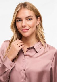 Satīna krekls ar apkakli mīkstā maigi rozā krāsā ar pogām priekšpusē, garām piedurknēm un gludu tekstūru, ko valkā modelis ar viļņainiem blondiem matiem.