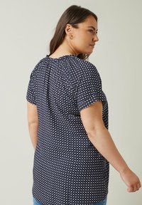 Marineblaue Bluse mit kurzen Ärmeln, gepunktetem Muster, gerafftem Ausschnitt und lockerer Passform, die von hinten eine glatte Stoffstruktur zeigt.