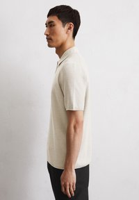 Beige short-sleeve polo shirt with a textured knit pattern, featuring a classic collar and a straight cut, paired with black trousers.