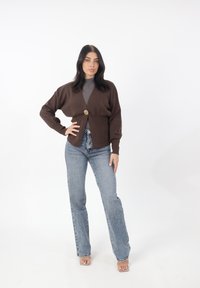 Cardigan marrón de punto con escote en V y cierre de un solo botón dorado, combinado con jeans de tiro alto en azul claro, sobre un fondo blanco.