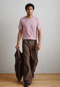 Ανοιχτό ροζ κοντομάνικο μπλουζάκι, χαλαρό καφέ παντελόνι φαρδιάς γραμμής και μαύρα παπούτσια. Το άτομο κρατά ένα σκούρο καφέ μπουφάν. Ορατές λείες υφές.