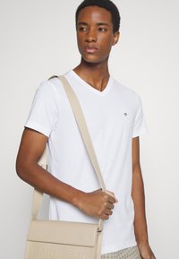 Jonge man die een wit V-hals T-shirt draagt en een beige schoudertas met reliëftekst bij zich heeft, kijkt iets naar de zijkant.