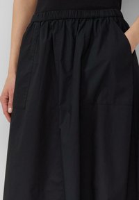 Detailní záběr na osobu v černé sukni s elastickým pasem a kapsami, jedna ruka odpočívá uvnitř kapsy.