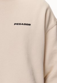Felpa in pile beige con finitura testurizzata, dotata di un logo nero "PEGADOR" nella parte superiore sinistra del petto.