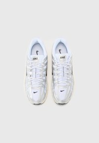 Bílé tenisky Nike mají síťovaný svršek se stříbrnými akcenty, černým logem a zavazovacím designem; polstrovaná podrážka je zobrazena z vrchu.