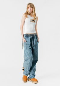 Top blanco sin mangas con un parche de pelaje, combinado con pantalones de denim azul holgados que tienen bolsillos laterales y una cinturilla elástica negra. Zuecos marrones.