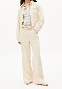 Beige kort jacka med en enda bröstficka, lager på lager över en blå och grå rutigt skjorta, i kombination med vida beige byxor.