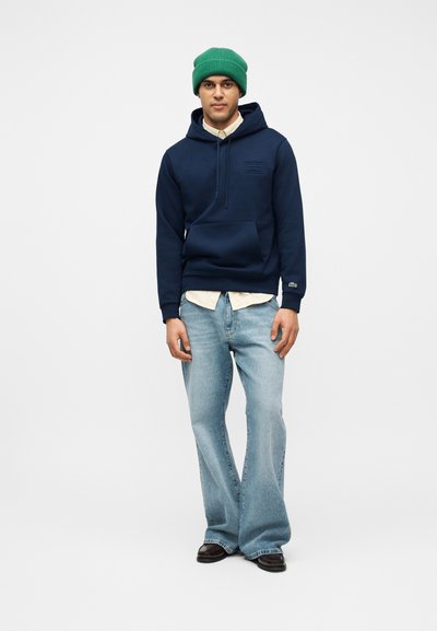 Homme portant un bonnet vert, un sweat à capuche bleu marine sur une chemise beige, un jean large bleu clair et des chaussures marron foncé, debout devant un fond blanc.