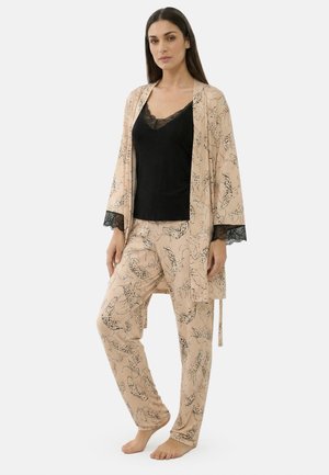 Donna in piedi a piedi nudi che indossa pantaloni del pigiama beige con motivo e vestaglia abbinata con rifiniture in pizzo nero sopra una canottiera con rifiniture in pizzo nero.
