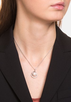 Collar de plata con un colgante circular que presenta una perla central, rodeada por un anillo texturizado. Llevado con un blazer negro.