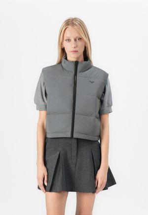 Vrouw in een grijze cropped puffer vest over een shirt met korte mouwen, gecombineerd met een hoogtaille donkergrijs plisséskirt, voor een effen achtergrond.