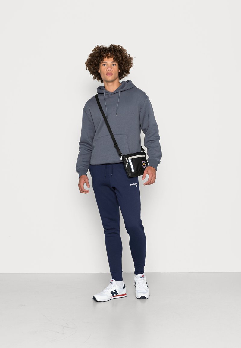 Un sweat à capuche gris avec une poche kangourou, un pantalon de sport bleu marine, et des baskets blanches avec des accents noirs et rouges. Petit sac à bandoulière noir.