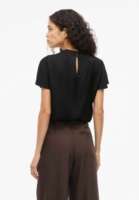 Blusa nera a maniche corte con dettaglio cut-out sulla schiena, morbida al tatto e dal fit ampio. Indossata con pantaloni marroni a vita alta.
