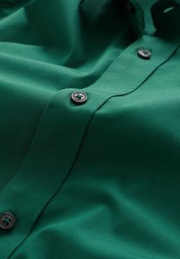 Grön skjorta med knappar framtill, som har en slät yta, mörka knappar med ett litet centrumhål och diskret sömnadsdetalj längs placket.