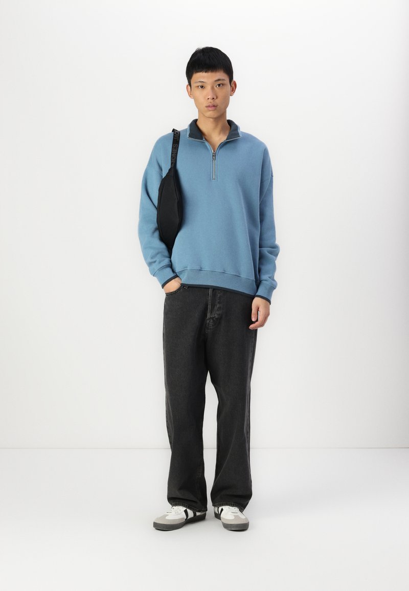 Blauwe fleece pullover met een half-rits kraag, gecombineerd met losse zwarte jeans en witte sneakers met zwarte accenten. Zwarte schoudertas.