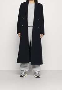 Abrigo largo de lana marino oscuro y de doble botonadura con grandes botones, llevado sobre un sudadera gris y pantalones de chándal, combinado con zapatillas multicolores.