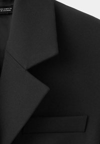Gros plan sur le revers et la poche passepoilée d'une veste noire ajustée, montrant la finesse de la texture du tissu et les détails des coutures.