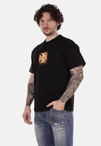 T-shirt in cotone nero con un design grafico rosso e oro sulla parte frontale con la parola "PROPAGANDA". La vestibilità è rilassata, adatta per un look informale.