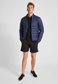 Marinblå pufferjacka med horisontell quiltning, full dragkedja, fickor på sidorna; matchas med en svart polo, svarta shorts och svarta sportskor.