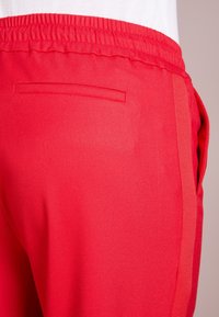 Röda, texturerade tygbyxor med elastiskt midjeband, sidofickor och en liten, horisontell bakficka.