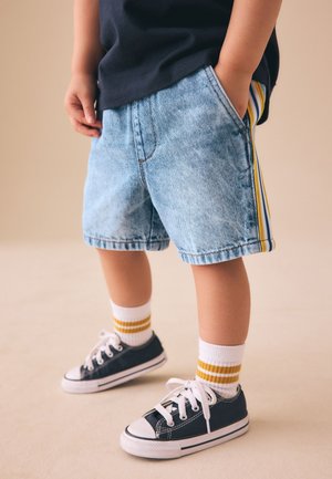 Kind trägt hellblaue Jeansshorts mit gelben Streifen an den Seiten, ein schwarzes Shirt, weiße Socken mit gelben Streifen und schwarze Sneakers.