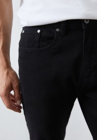 Hombre con pantalones de mezclilla negros ajustados y una camisa blanca metida por dentro, con una mano relajada visible a un lado.