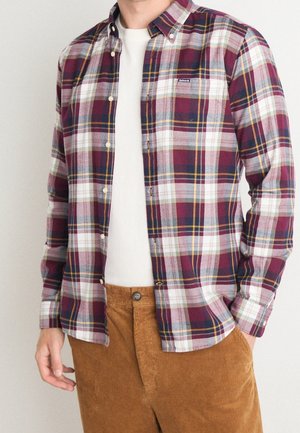 Mężczyzna ubrany w rozpiętą bordowo-biało-granatową koszulę w kratę na białej koszulce, noszący brązowe spodnie sztruksowe.