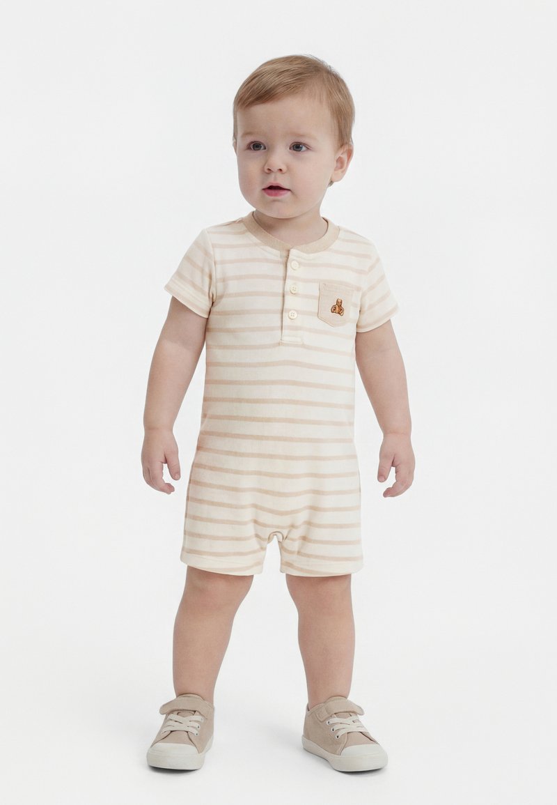 Copil mic stă în picioare, purtând un salopetă bej și albă în dungi cu nasturi și buzunar, și adidași bej, pe fundal alb simplu.