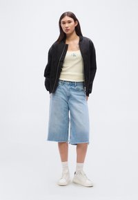 Μαύρο μπουφάν τύπου bomber πάνω από ανοιχτό κίτρινο τοπ με μικρό λογότυπο, σετ με φαρδιά ανοιχτό μπλε τζιν σορτς και λευκά αθλητικά παπούτσια.