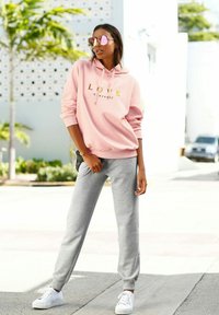 Lyserød hoodie med gylden tekst "ELSK dig selv," parret med grå sweatpants og hvide sneakers. Spejlsolbriller fuldender looket.