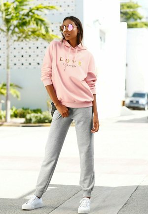 Lichtroze hoodie met gouden tekst "LOVE yourself," gecombineerd met grijze sweatpants en witte sneakers. Spiegelzonnebril maakt de outfit compleet.