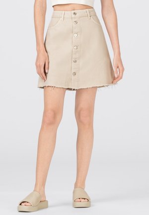 Kvinde iført en beige denim-mini nederdel med knapper foran og frynset kant samt beige slip-on platformsandaler, stående foran en ensfarvet baggrund.