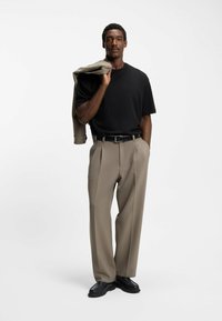 Uomo con una maglietta nera e pantaloni beige che tiene una giacca beige abbinata sulla spalla, in piedi davanti a uno sfondo bianco uniforme.