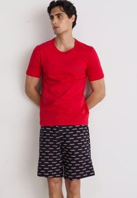 Rotes Baumwoll-T-Shirt mit Rundhalsausschnitt und kurzen Ärmeln, kombiniert mit schwarzen Shorts, die ein wiederholtes rotes Logo-Muster aufweisen.