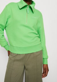 Zelený svetr s vysokým límcem a půlzipem, se strukturovaným povrchem a přiléhavým pasem, doplněný olivovými kalhotami s širokými nohavicemi.