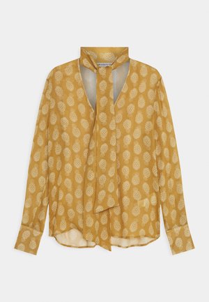 Musztardowo-żółta bluzka na długi rękaw z białym wzorem ananasów i ozdobnym wiązaniem przy szyi.