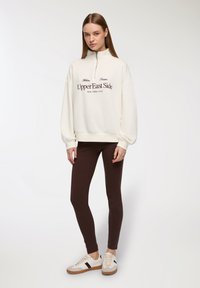 Biała bluza zapinana na zamek z napisem "Upper East Side", połączona z brązowymi legginsami i beżowymi trampkami z czarnymi akcentami.