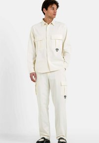 Camisa de botón color crema con dos bolsillos en el pecho y un acento de logo triangular, combinada con pantalones cargo a juego que tienen bolsillos laterales.