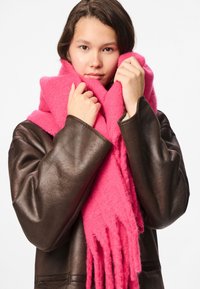 Lyserød uldtørklæde med frynser, lagt over en mørkebrun læderjakke. Blød tekstur tilføjer varme og dybde.