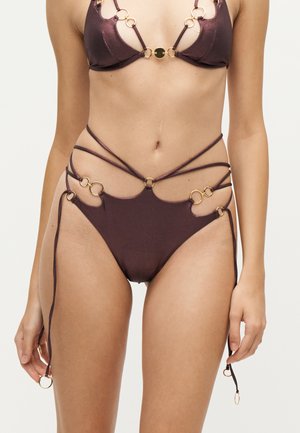 Bikini marron avec des détails en anneaux dorés et plusieurs fines bretelles autour de la taille et de la poitrine sur une personne debout.