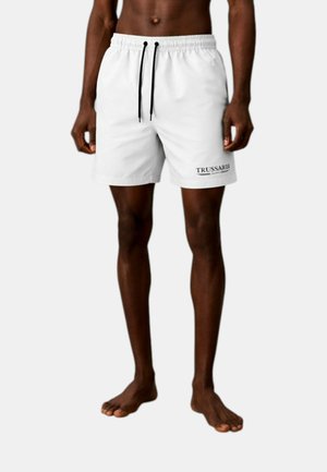 Uomo che indossa pantaloncini bianchi con coulisse e logo Trussardi sulla gamba sinistra, in piedi a piedi nudi su uno sfondo chiaro e semplice.