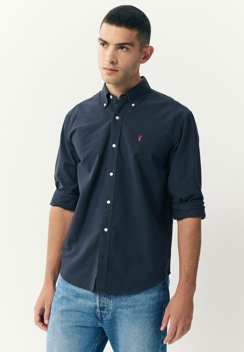Joven con una camisa de botones azul marino con mangas arremangadas y jeans azul claro, mirando ligeramente de lado contra un fondo sencillo.