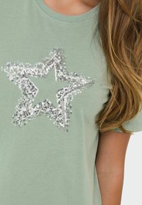 Lys grøn bomulds T-shirt med et sølvfarvet palietstjerne design i midten. Kortærmet og blød tekstur med rund halsudskæring.