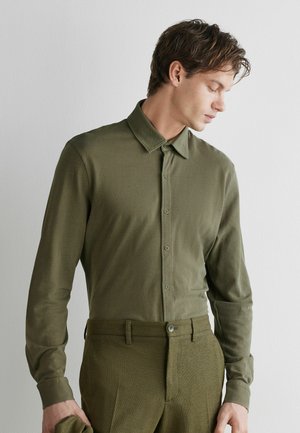 Jeune homme portant une chemise à manches longues boutonnée vert olive et un pantalon assorti, regardant vers le bas et tenant un tissu dans sa main gauche.