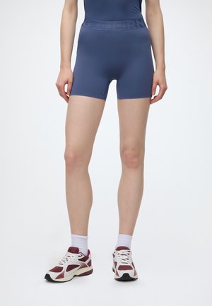 Personne portant un short bleu marine ajusté, des chaussettes blanches mi-mollet et des chaussures de sport bordeaux et blanches, debout sur un fond blanc.